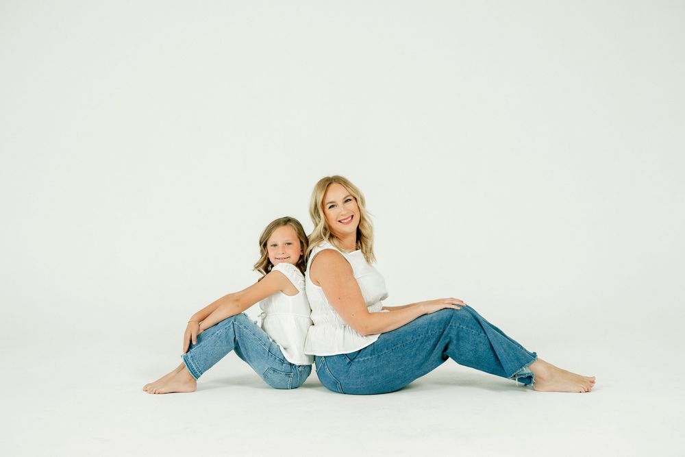 Two females in white tops and jeans sit back-to-back on a white studio floor, smiling at the camera.