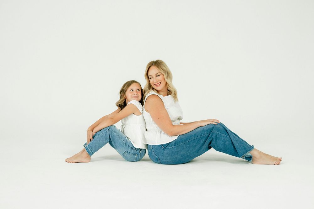 Two females in matching white tops and jeans sit back-to-back on a white studio floor, smiling warmly.
