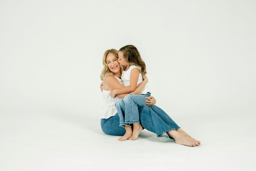 A mother and daughter in white tops and jeans sit together on a white background, sharing a tender hug and kiss.