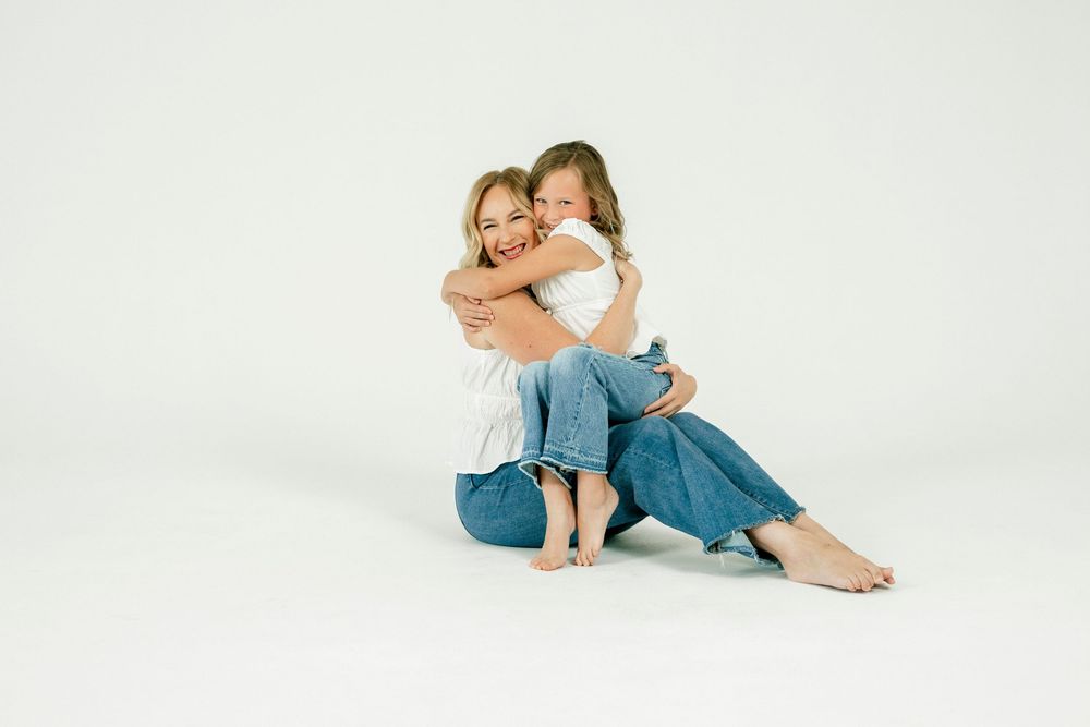 A mother and daughter in matching white tees and jeans share a warm hug while sitting on a white background.
