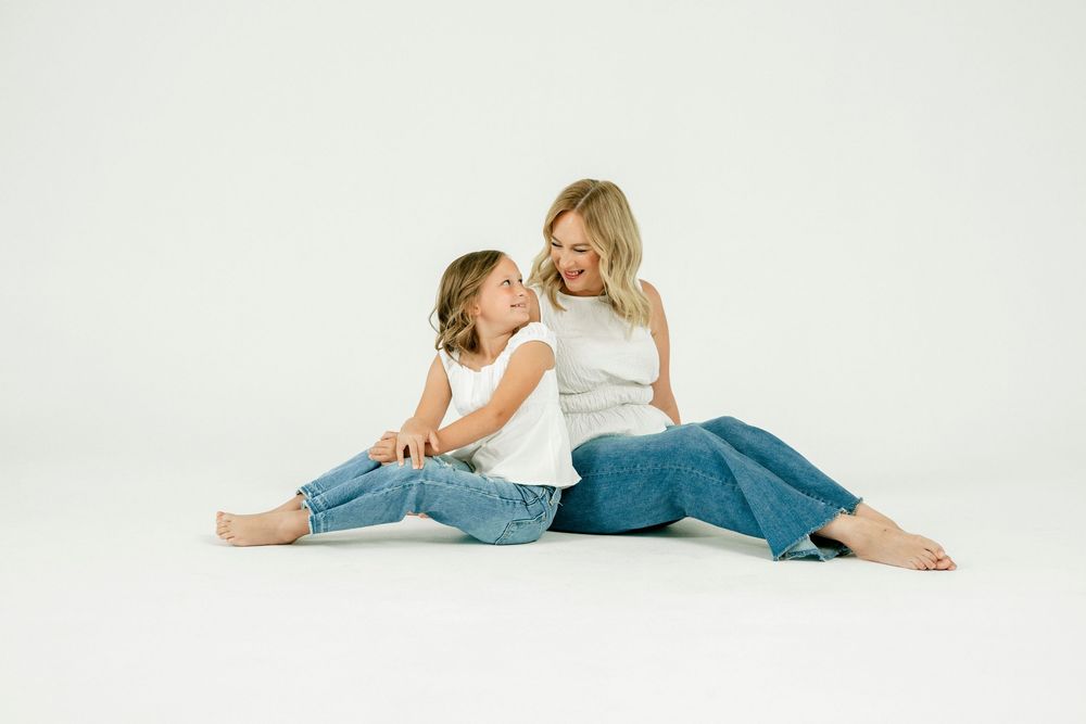 A woman and young girl in matching white tops and jeans sit together laughing on a white background.