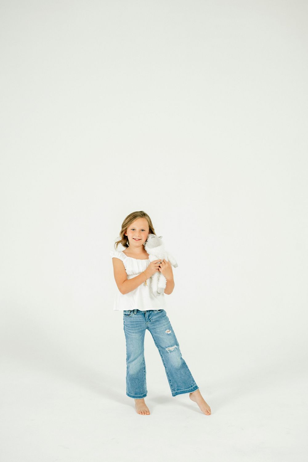 A smiling girl in white top and ripped jeans holds a stuffed animal toy while standing barefoot on white background.