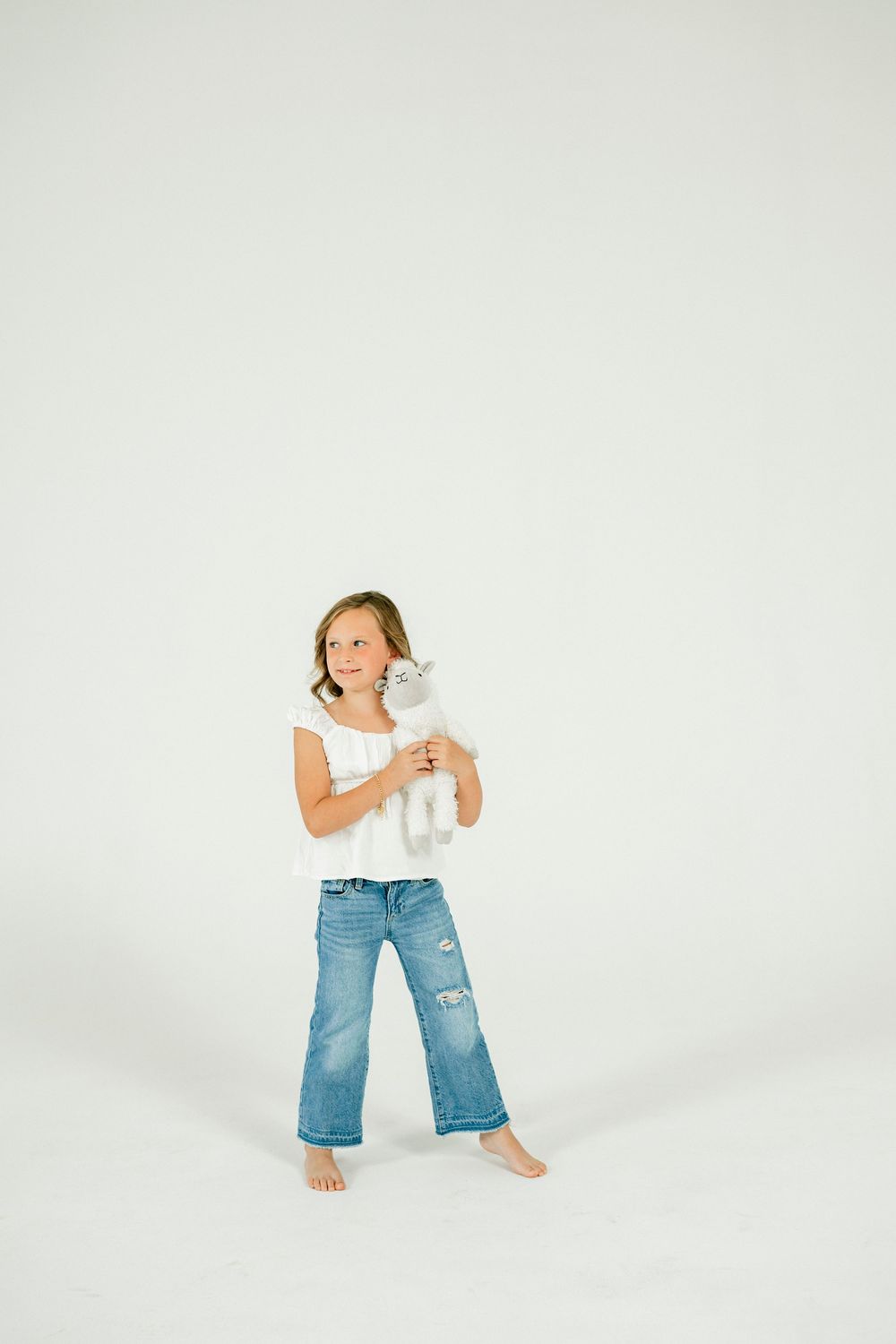 Young girl in white top and jeans holds a stuffed animal toy, smiling in a bright white studio setting.