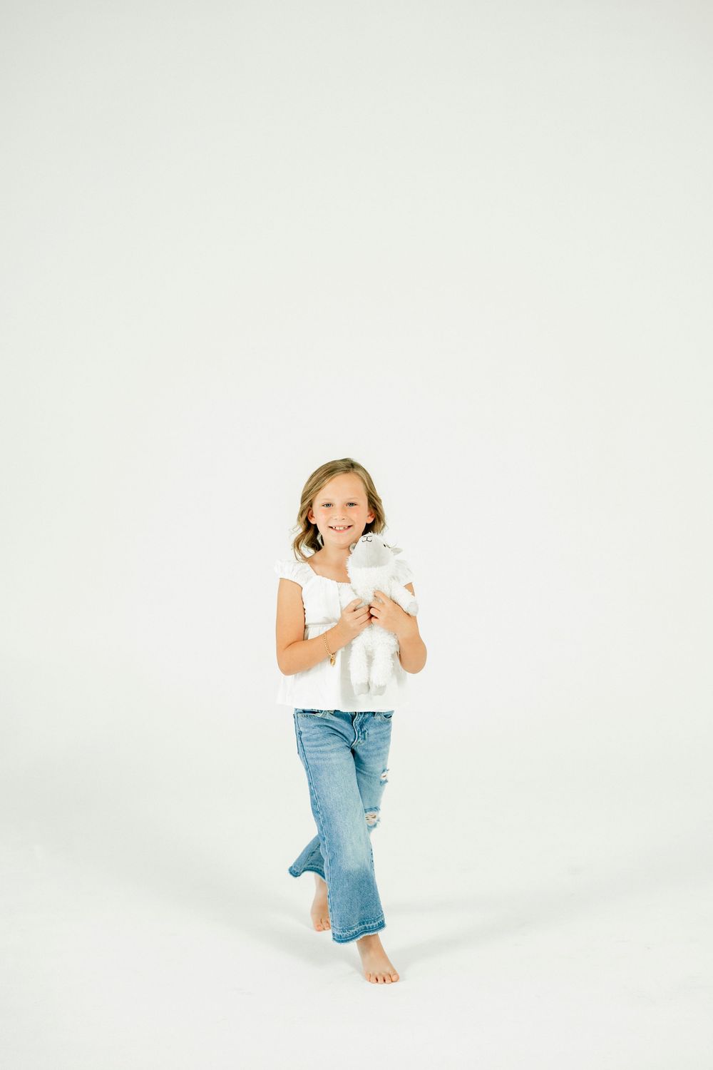 A smiling young girl in white tee and jeans holds a stuffed animal toy while standing barefoot on white background.