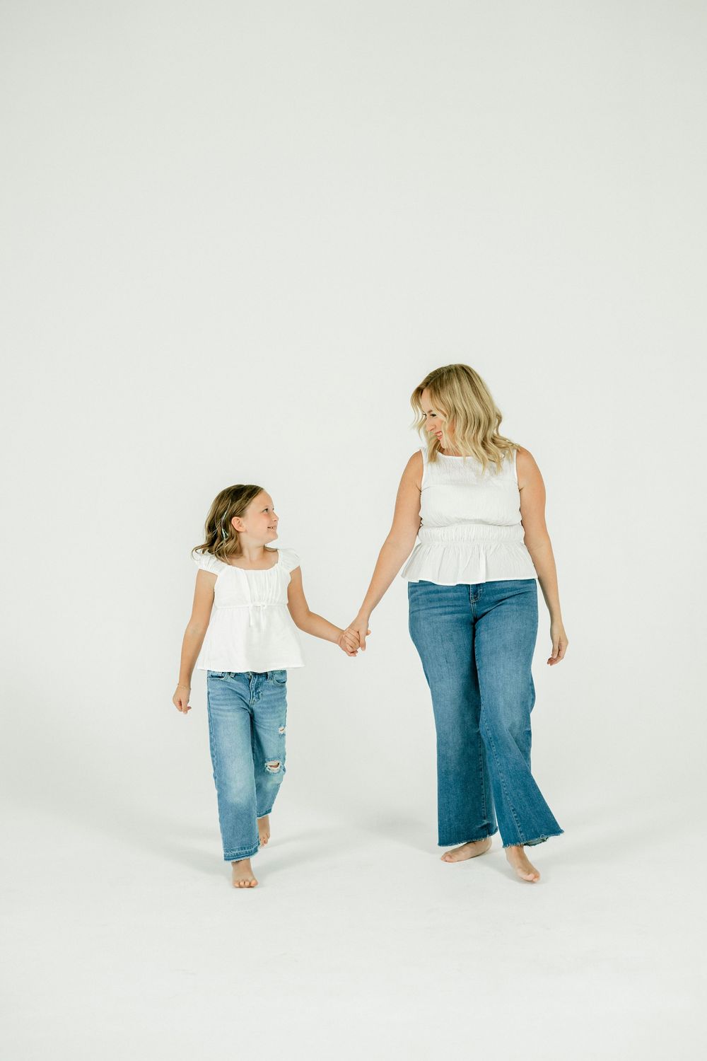 Mother and daughter in matching white tops and jeans hold hands walking barefoot on white background.