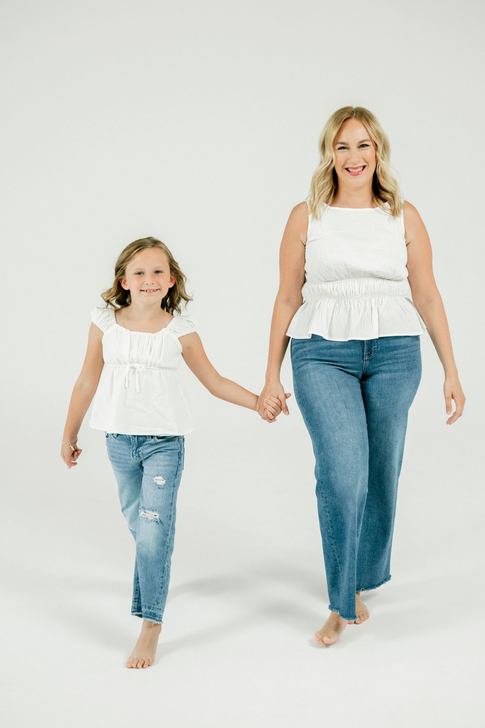 Smiling mother and daughter in white tops and jeans walk hand in hand barefoot on white background.