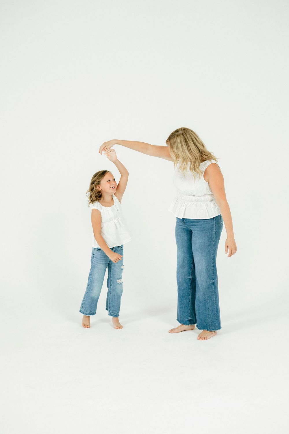 A mother and daughter in matching white tops and jeans dancing together on a white background.