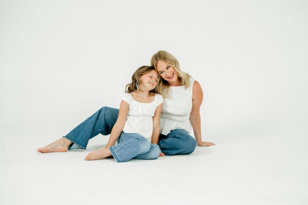 A woman and young girl in matching white tops and jeans sit together smiling on a white studio floor.
