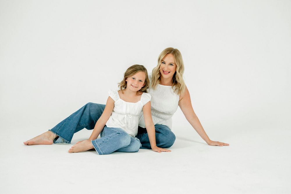 A woman and young girl in white tops and jeans sit together smiling on a white studio floor.