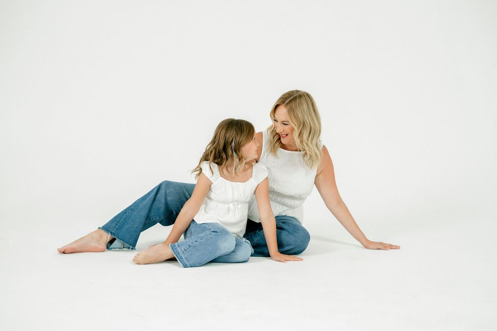 A blonde woman and young girl in white tops and jeans sit facing each other laughing on a white studio floor.
