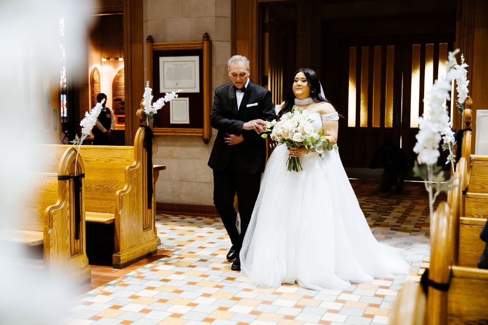 Bride in white gown walks down church aisle with escort, holding bouquet, surrounded by elegant floral decorations.