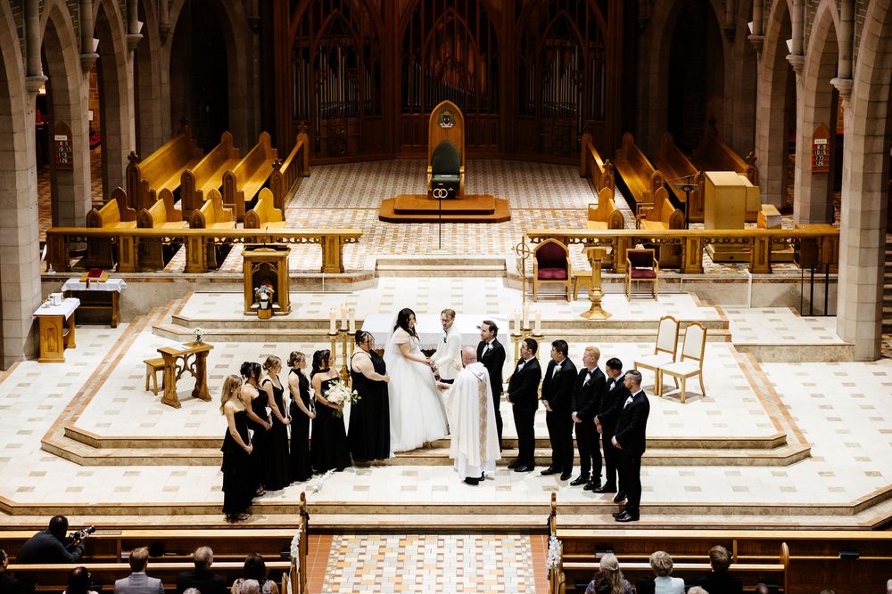 Elegant church wedding ceremony with bride, groom, bridesmaids in black, and groomsmen at ornate altar.