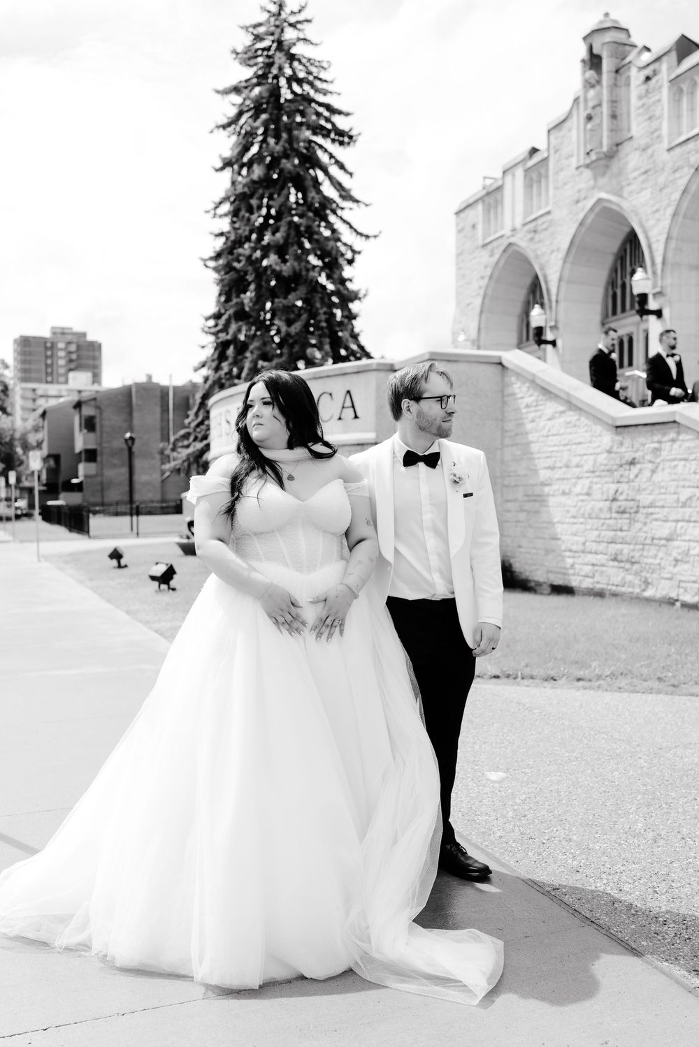 Black and white photo of bride and groom posing outside stone church beside tall evergreen tree.