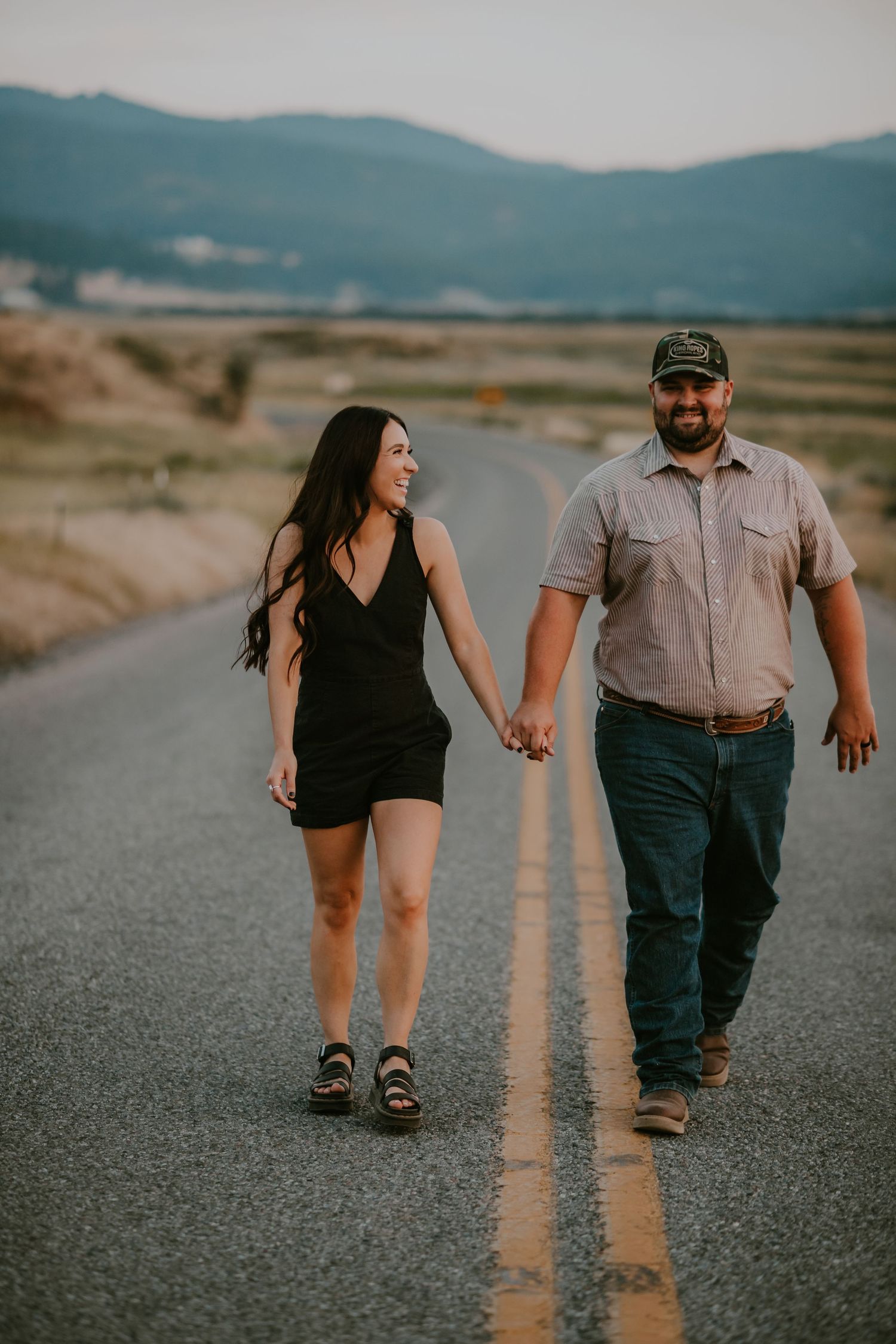 Couple holding hands walking down a rural road with mountains in the background during golden hour.