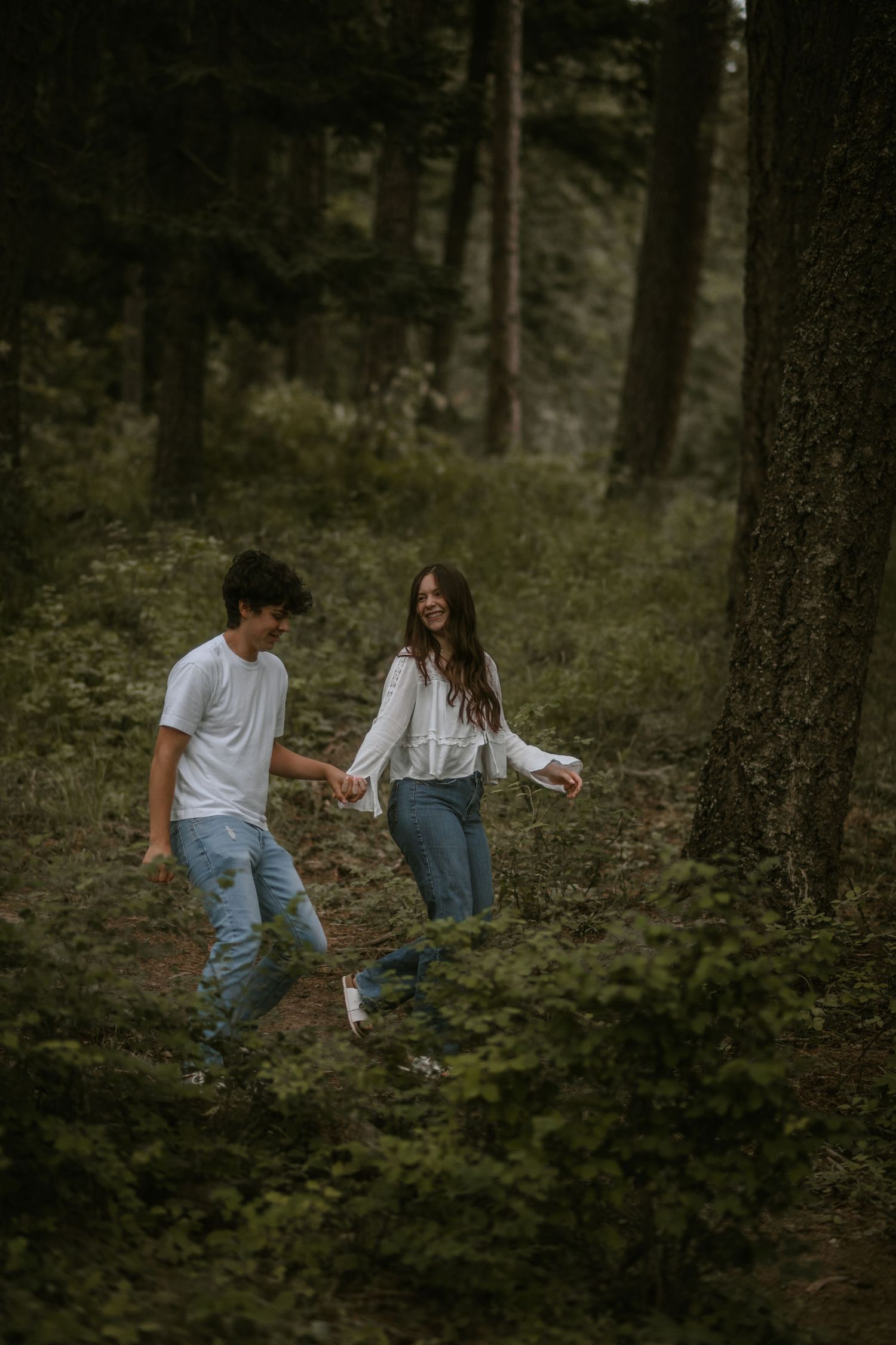 A young couple holds hands and laughs while walking through a lush green forest wearing white tops and blue jeans.