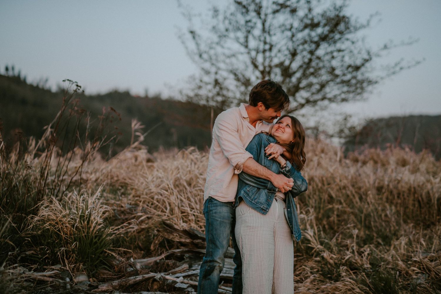 Couple embracing romantically in a golden field at dusk, man kissing woman's forehead near a bare tree.