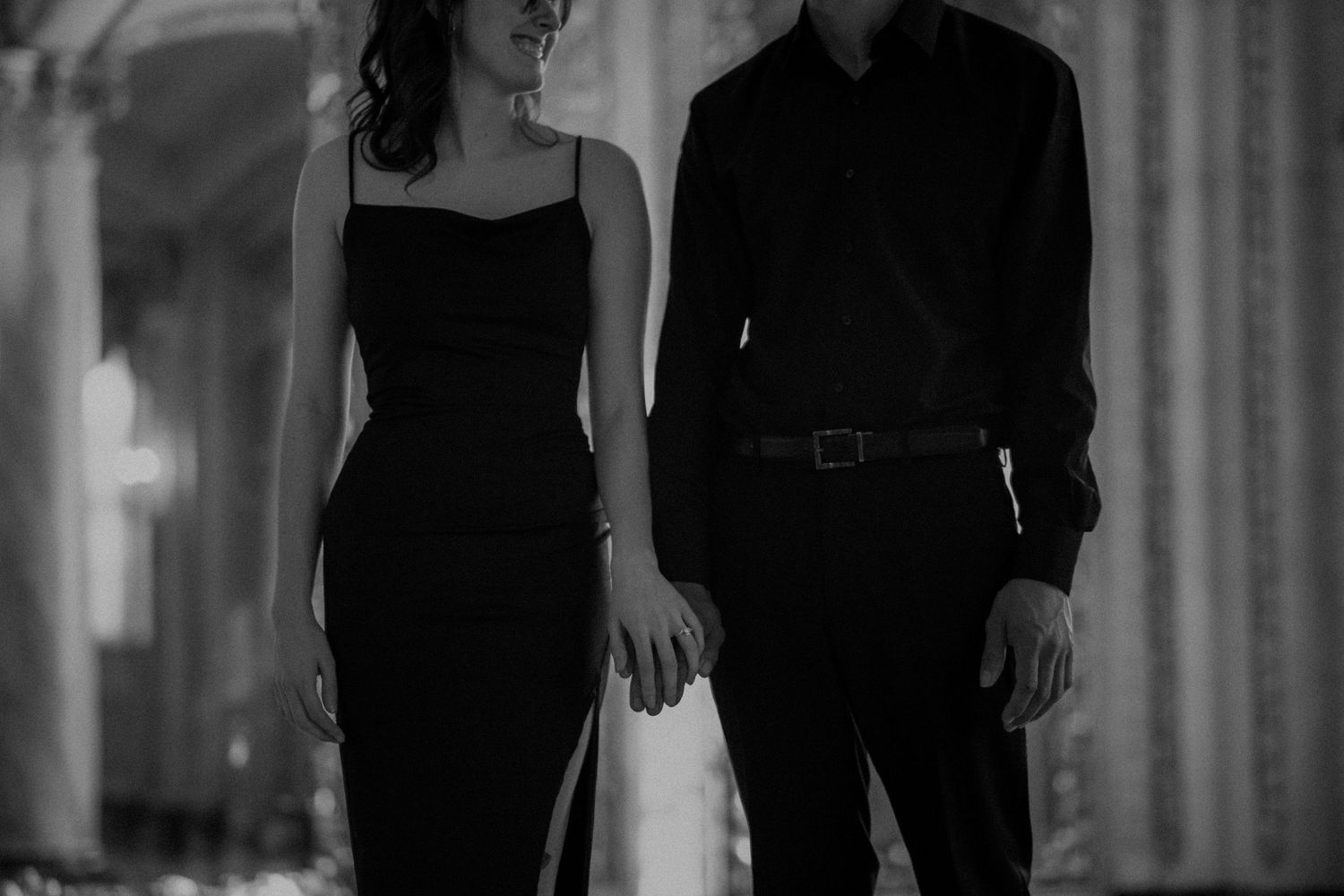 Elegant couple in matching black outfits holding hands in a classic architectural setting, black and white photo.