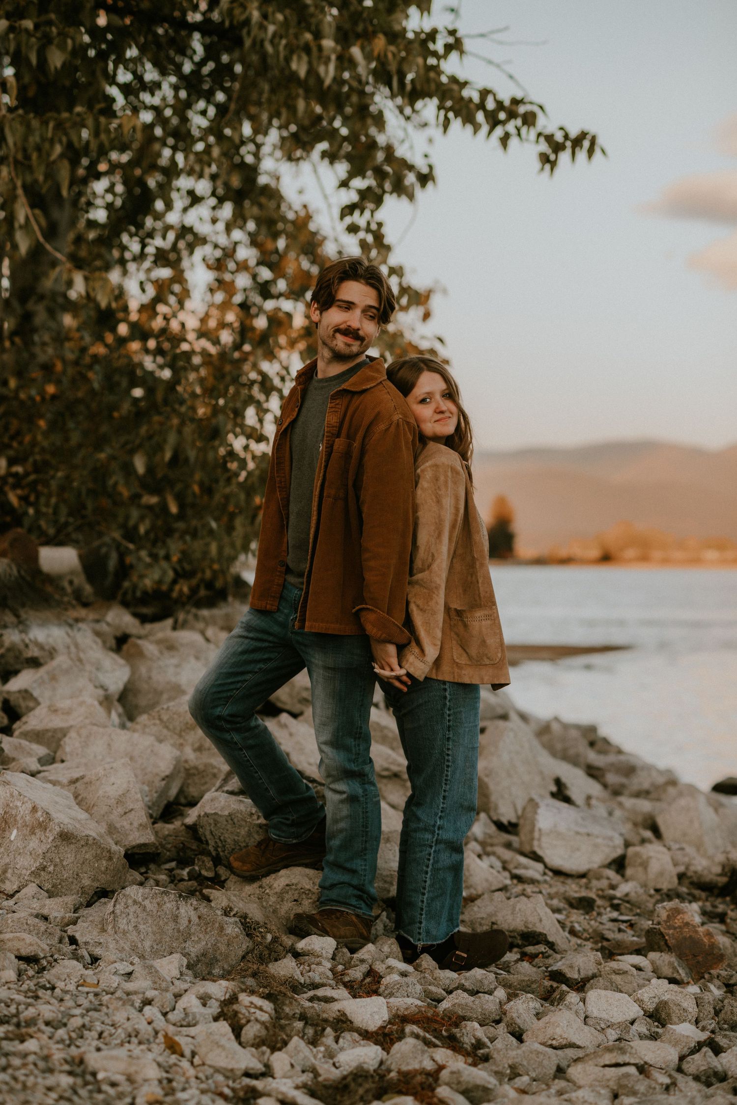 Couple in brown jackets and jeans holding hands on rocky lakeside shore with autumn trees and mountains at sunset.