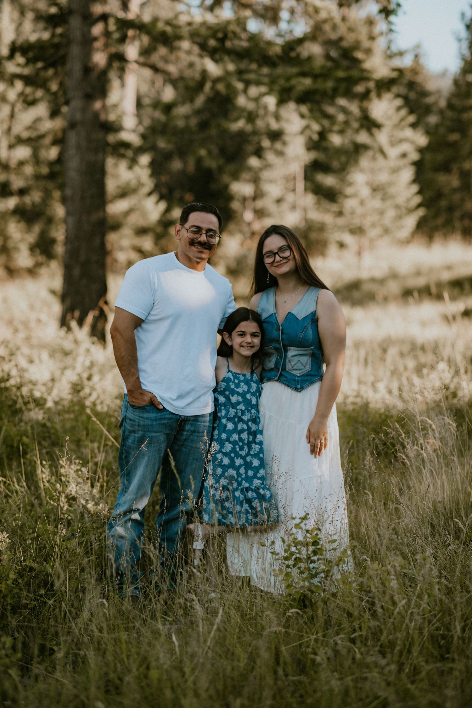A family of three poses in a sunlit meadow surrounded by tall grass and pine trees, wearing casual blue and white outfits.