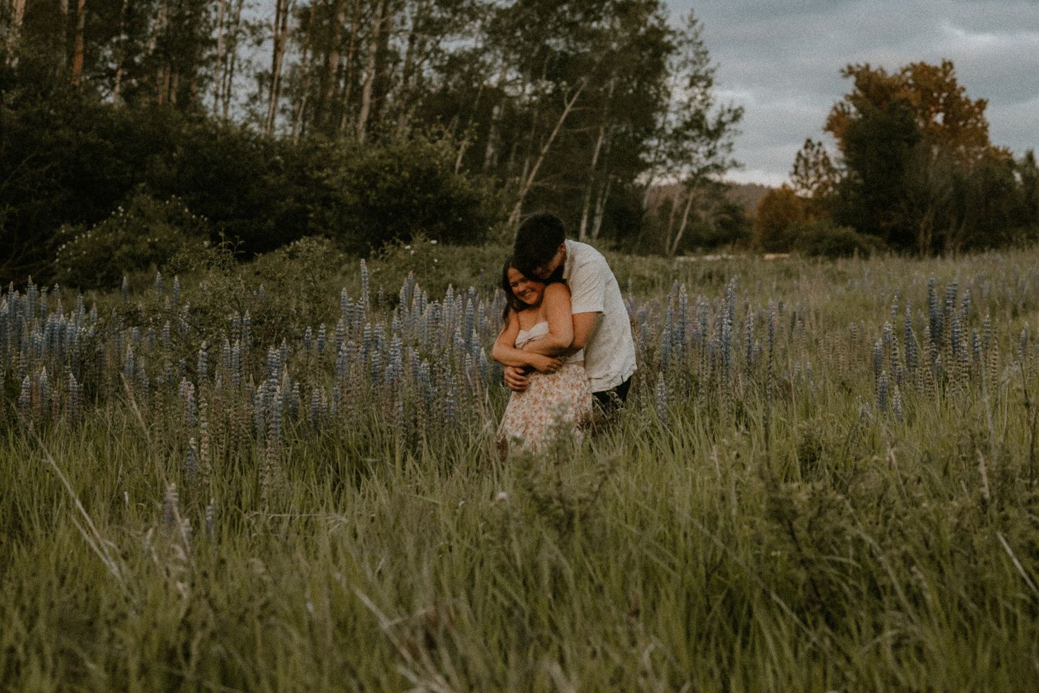 Couple embracing in a wildflower field with tall grass and trees at dusk.
