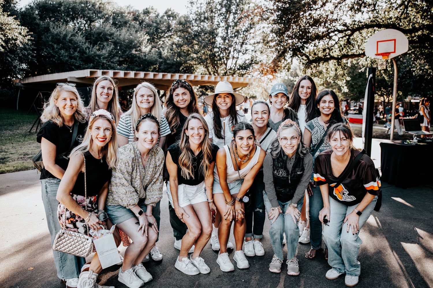 A large group of smiling young women posing together outdoors near a basketball court on a sunny day.