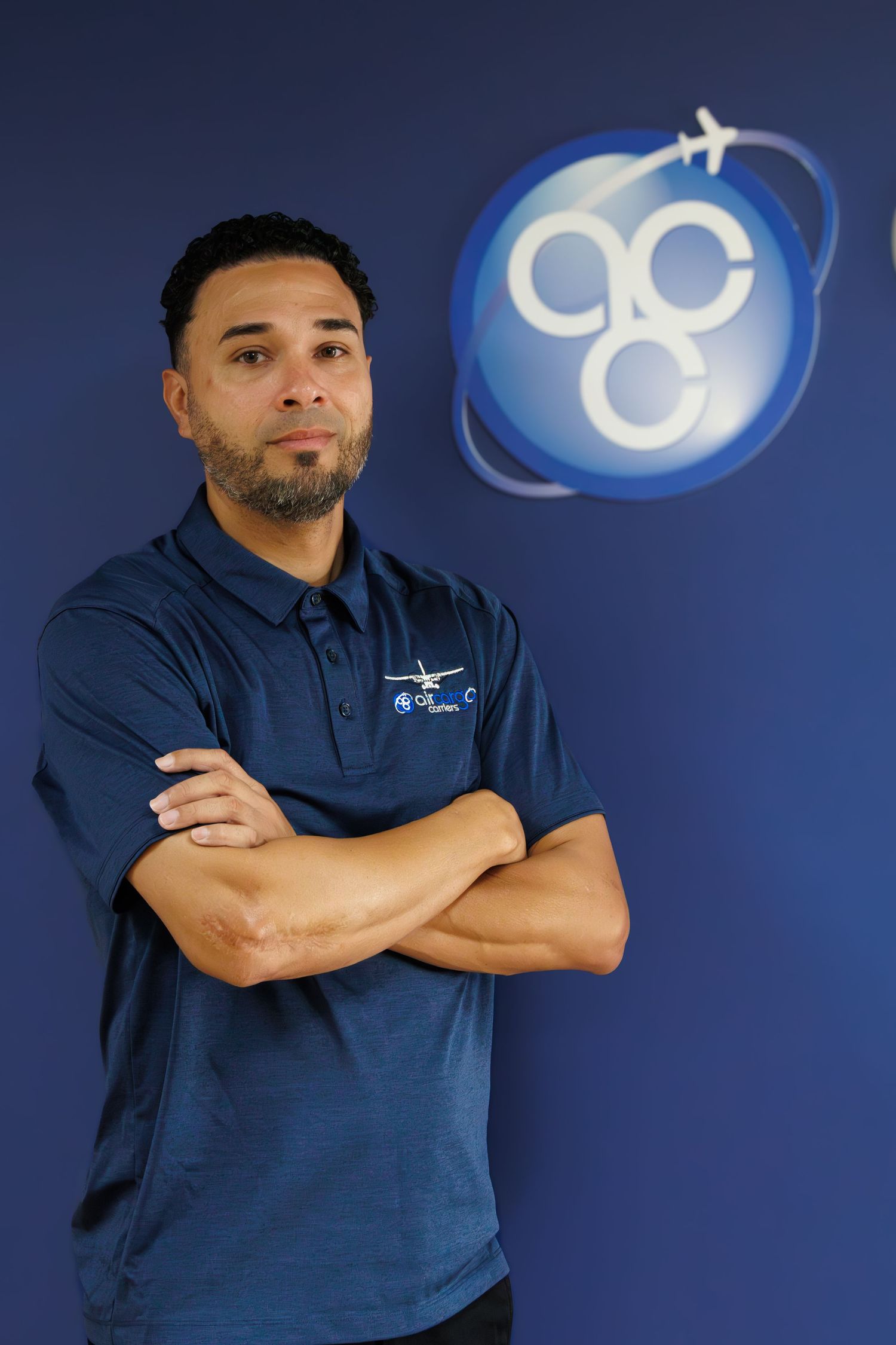 Man in navy polo shirt with arms crossed stands before blue wall with circular aviation company logo.