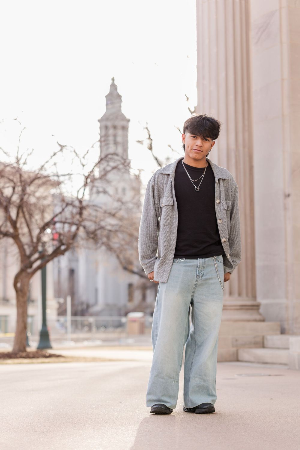 Teen in gray overshirt, black tee, and wide-leg jeans stands near classical building columns.