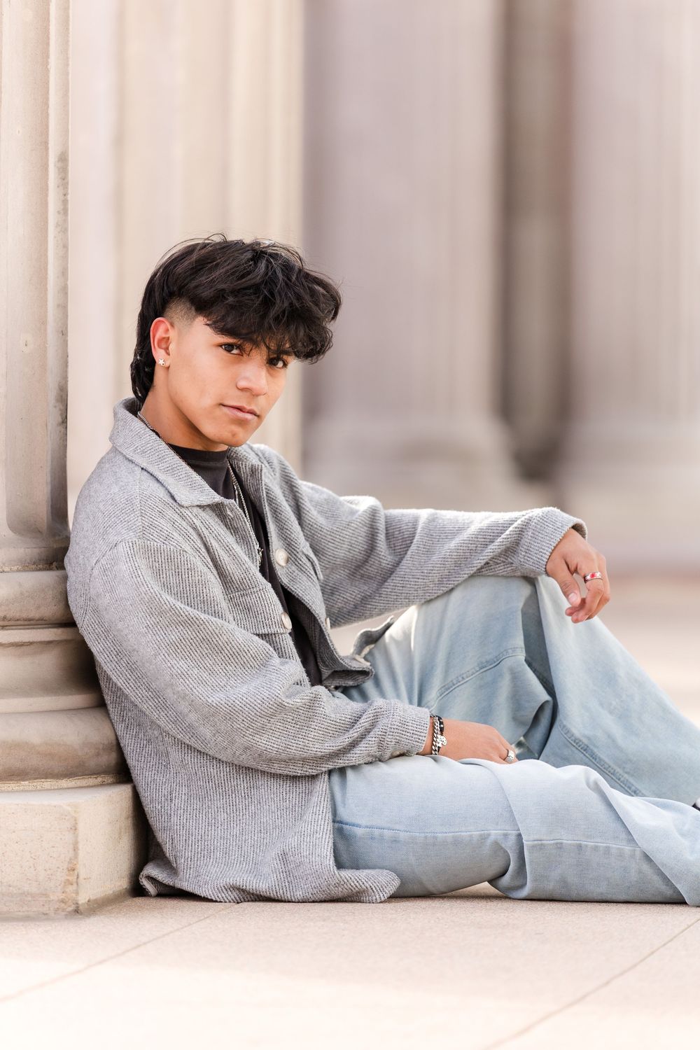 Stylish young man in gray jacket and jeans leaning against a stone column outdoors.