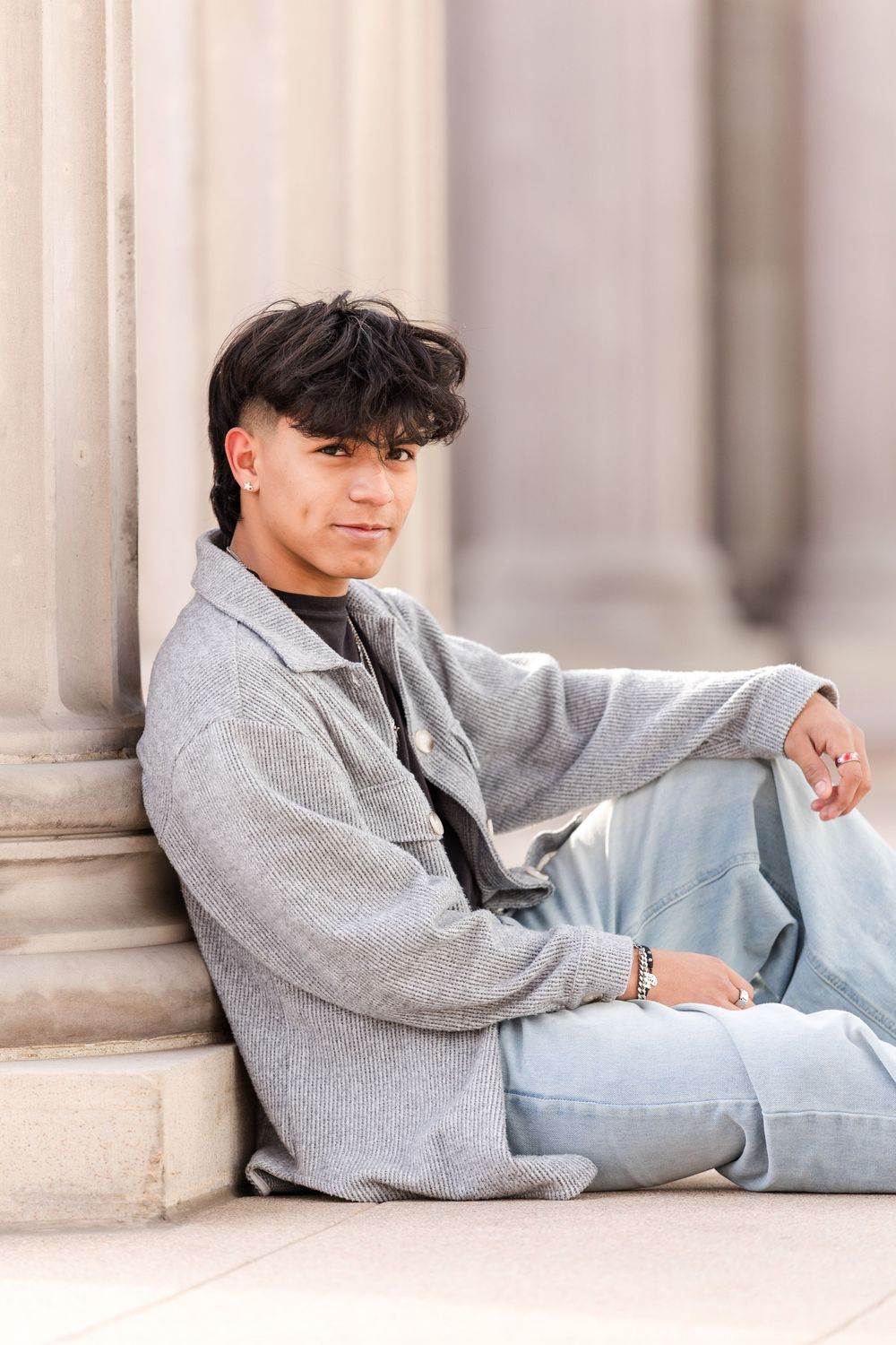 Young man in gray jacket and jeans sits relaxed against stone pillar outdoors.