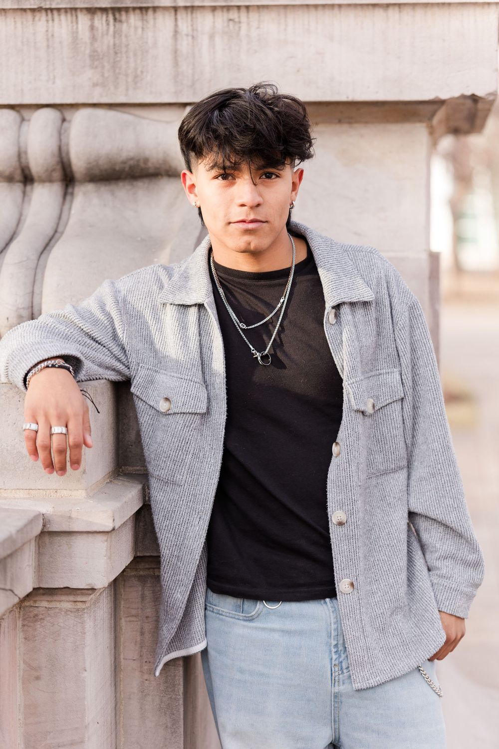 Young man in gray jacket and black shirt leaning against stone pillar outdoors.
