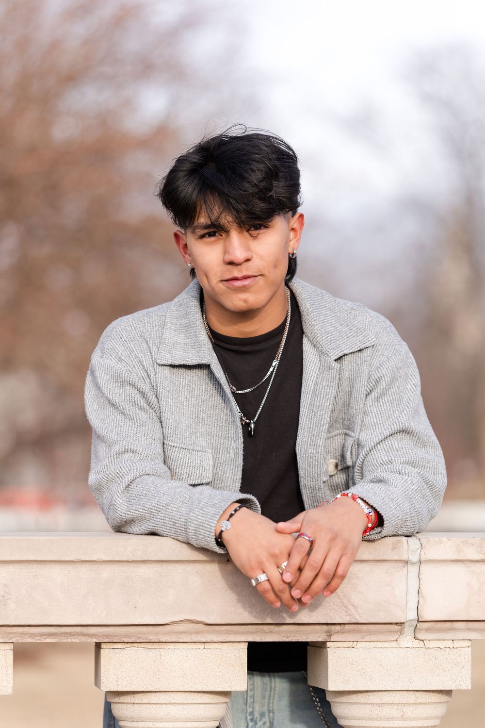 Young man in gray jacket leaning on stone railing outdoors, wearing silver necklace and ring, with blurred trees behind.