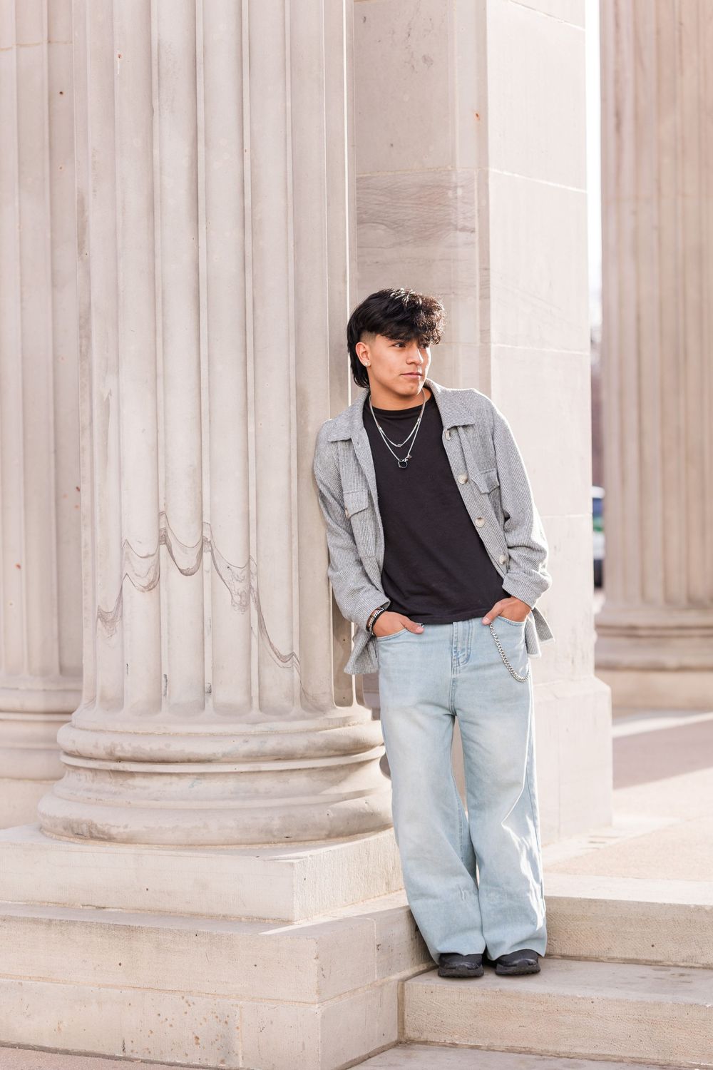 Young man in gray jacket and light jeans leaning against classical stone columns on steps.
