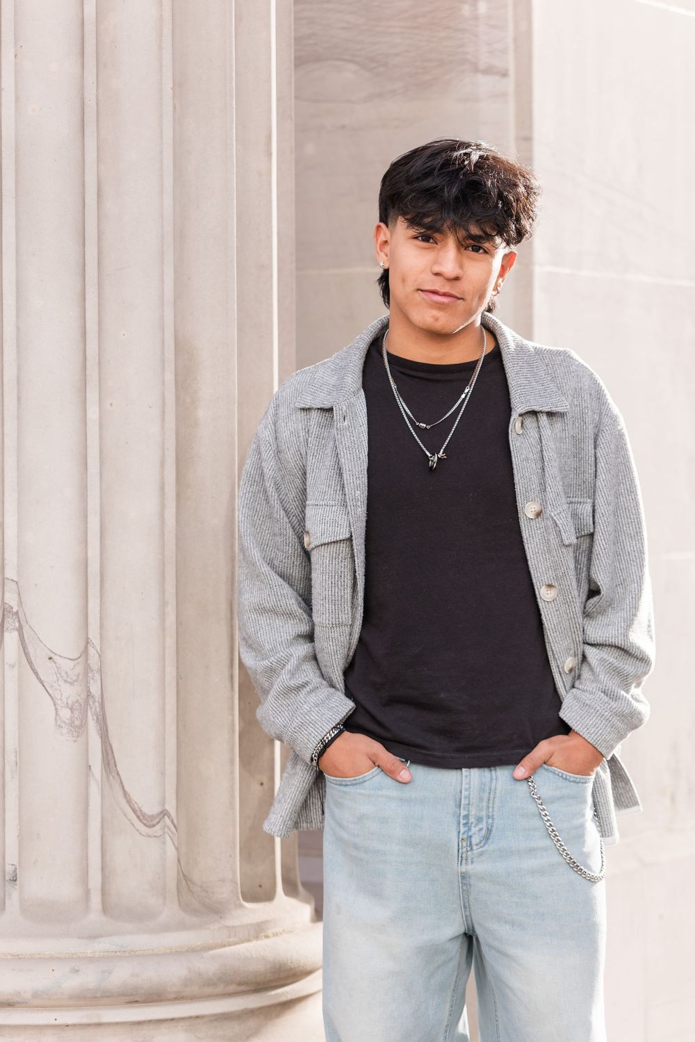 Young man in gray jacket, black tee, and light jeans poses against stone column.