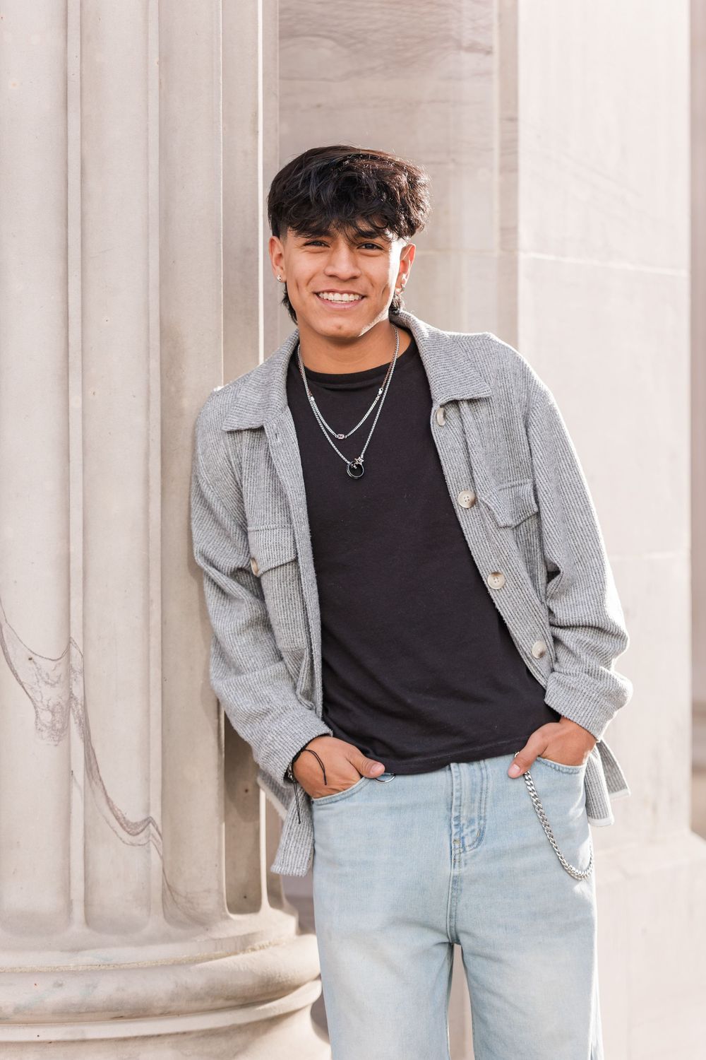 Smiling young man in gray jacket, black tee, and light jeans posing by stone columns.