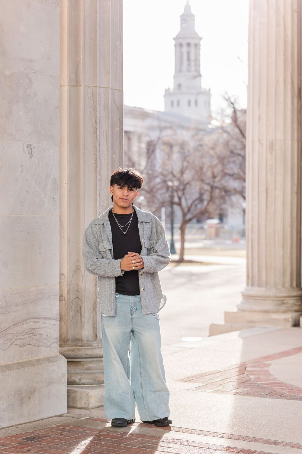 Young man in wide-leg jeans and denim jacket poses between columns of a classical building with tower backdrop.