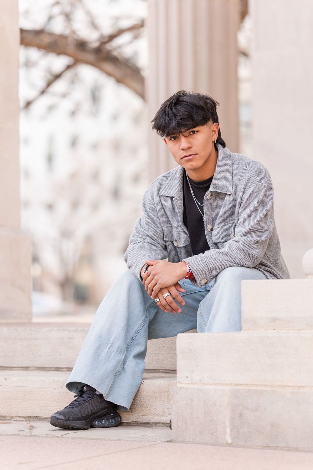 Young man in gray jacket and light jeans sitting on stone steps near columns.