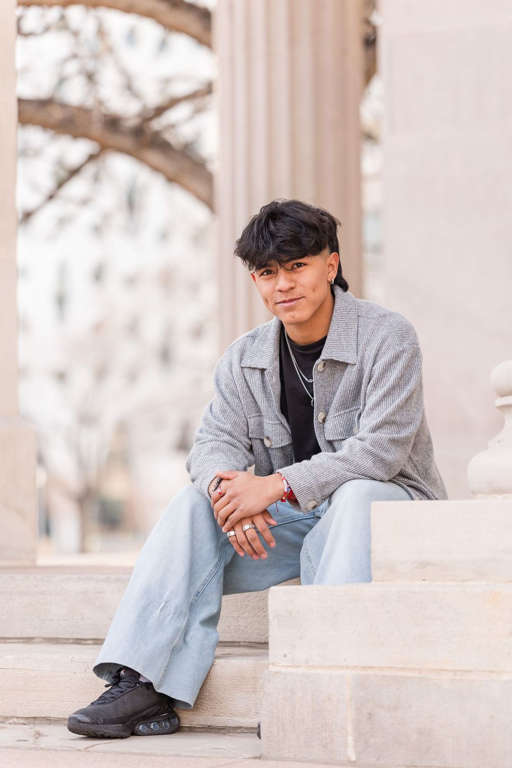 Young man in gray jacket and light jeans sitting on marble steps outdoors.