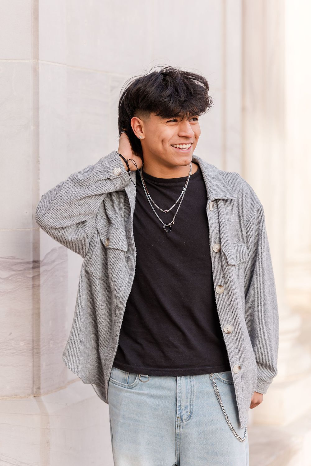 Smiling young man in gray overshirt and black tee leaning against stone wall.
