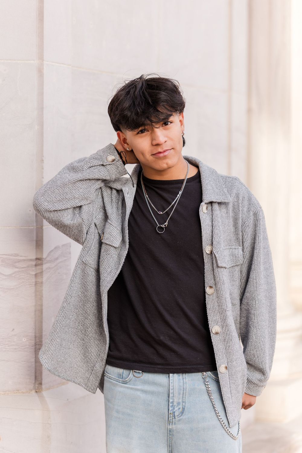Young man in gray shacket over black tee and jeans, wearing silver chain necklaces, leaning against a white wall.