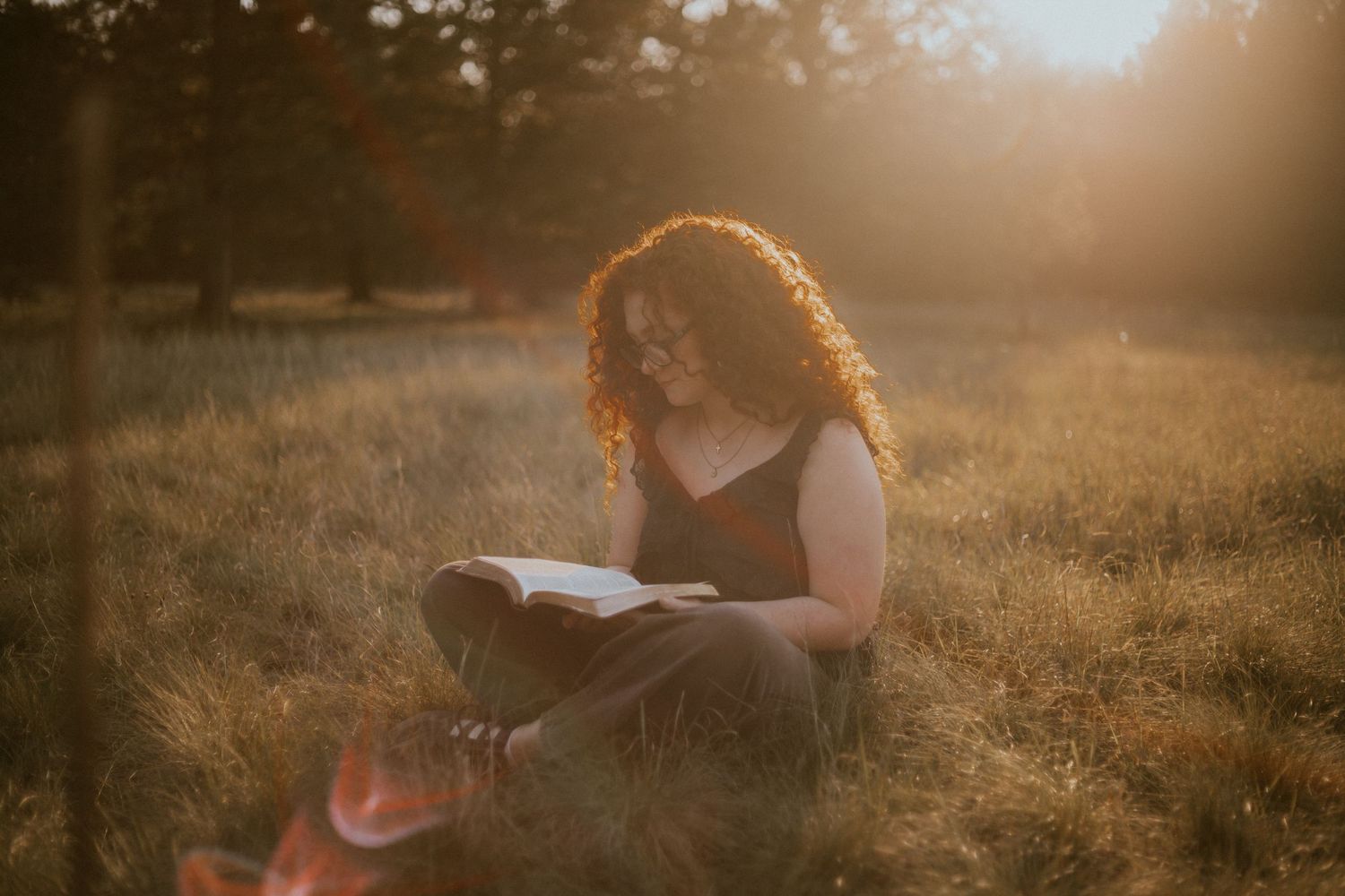 Woman with curly hair reading a book while sitting cross-legged in a sunlit meadow during golden hour.