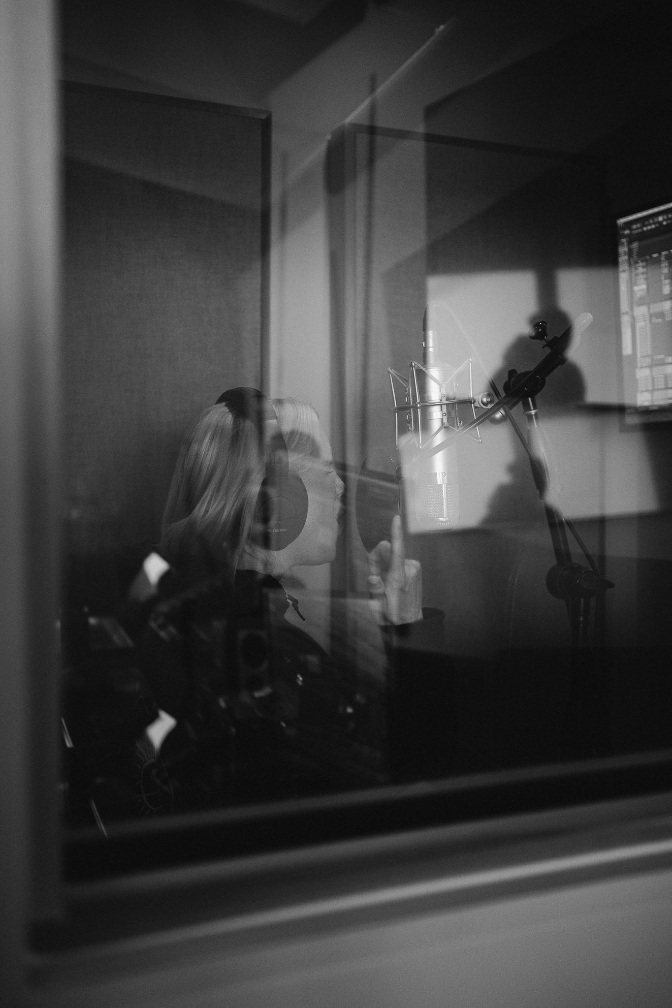 Black and white photo of a blonde woman recording vocals in a professional music studio booth with a condenser microphone.