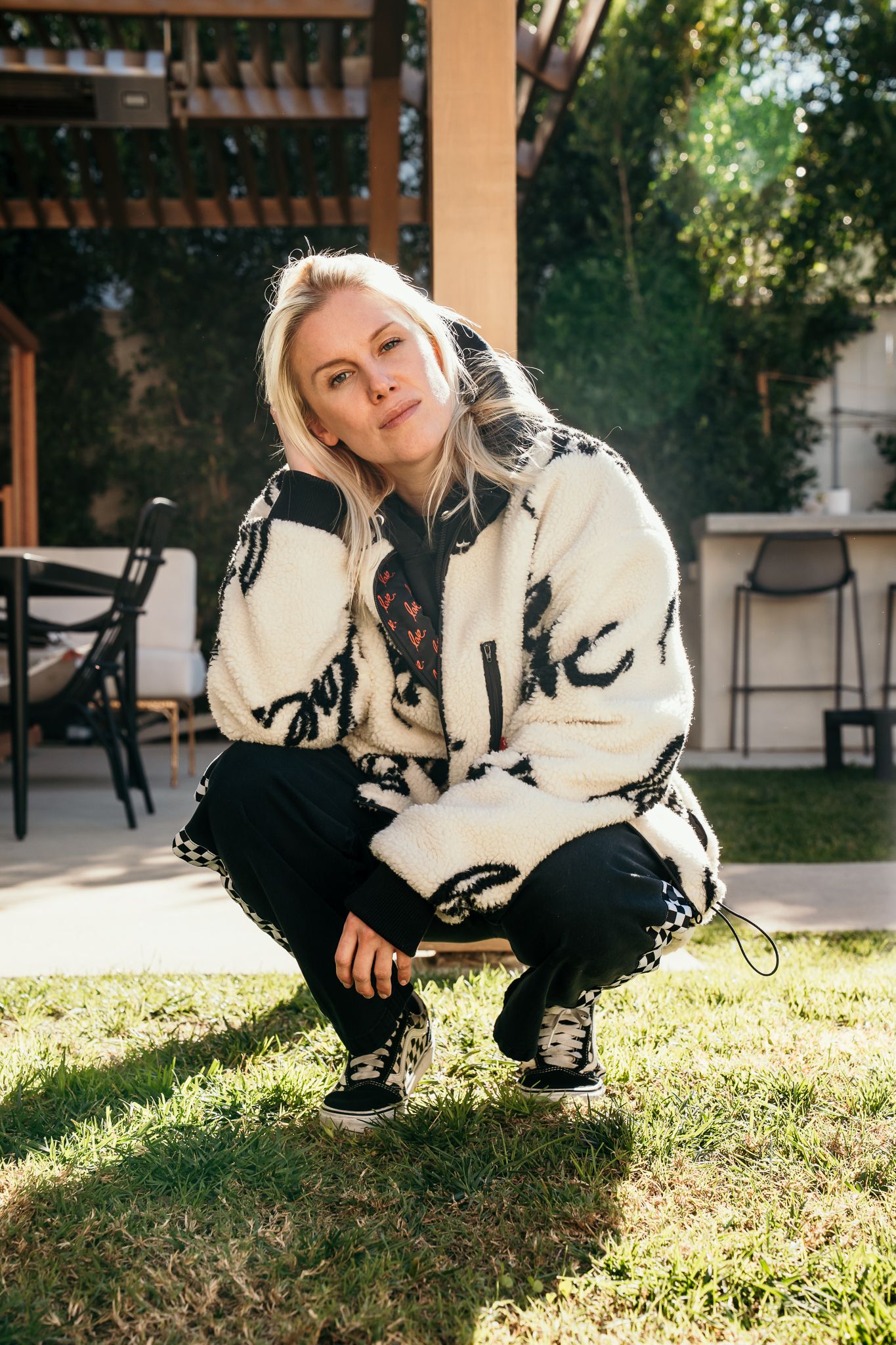 Blonde woman crouching on grass in a patterned white jacket and black pants, posing outdoors near a pergola.