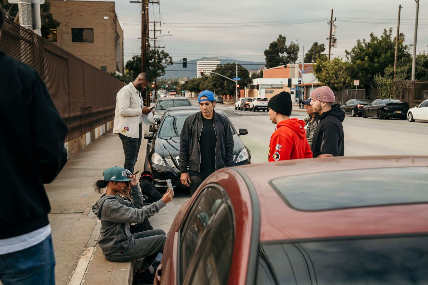 Group of people gathered on a city sidewalk near parked cars on a busy urban street.