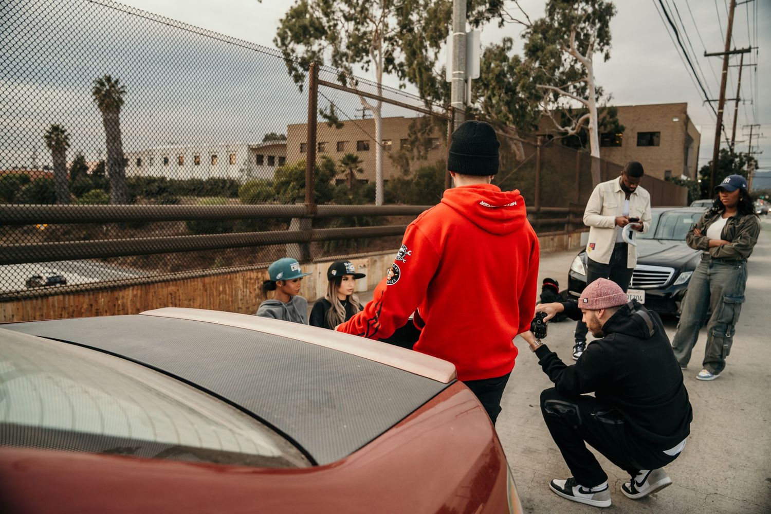 Group of young people gathered around a red car on an urban street near a chain-link fence in Los Angeles.