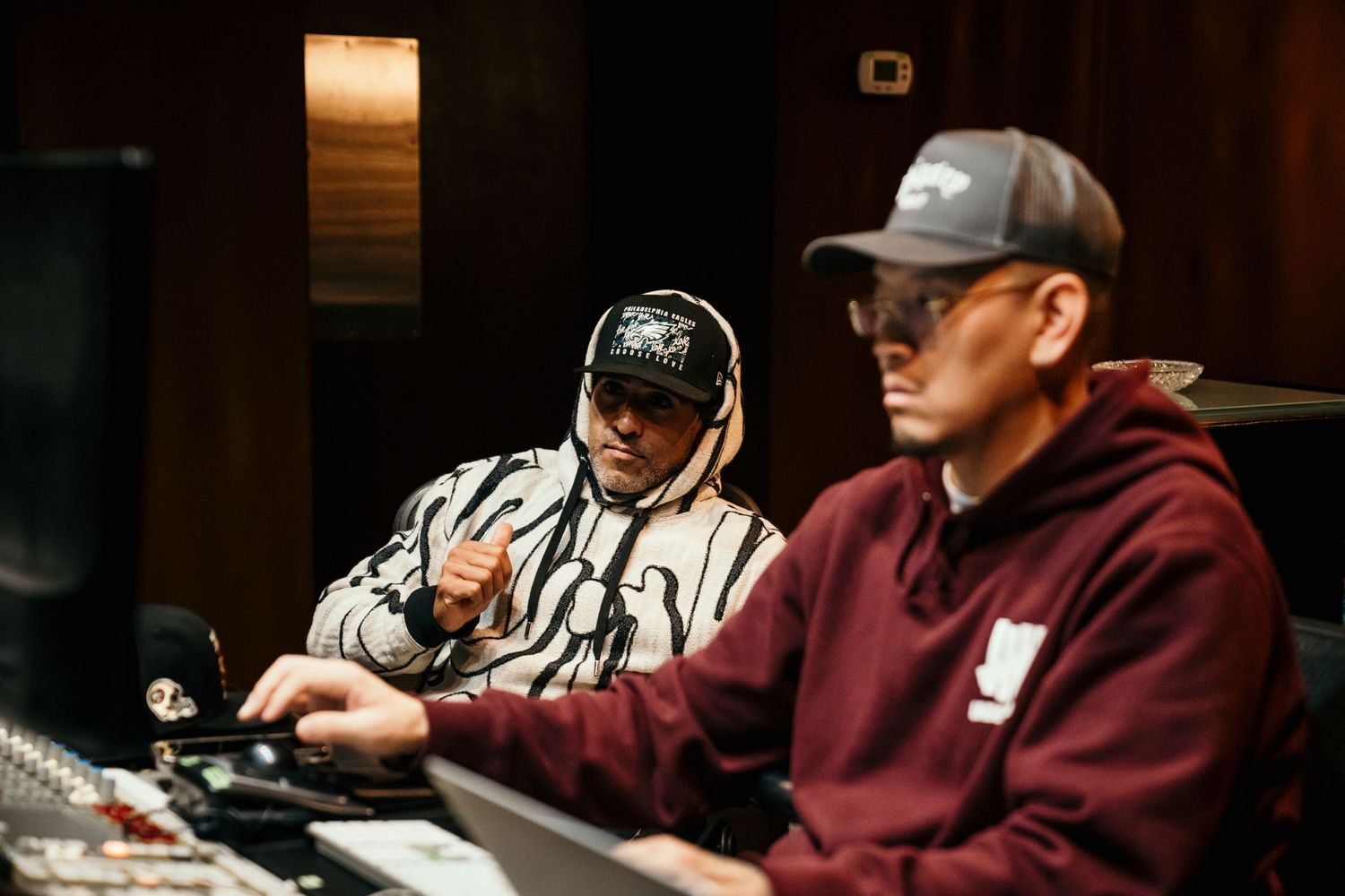 Two men wearing caps work at a professional music studio mixing console in a dimly lit recording session.