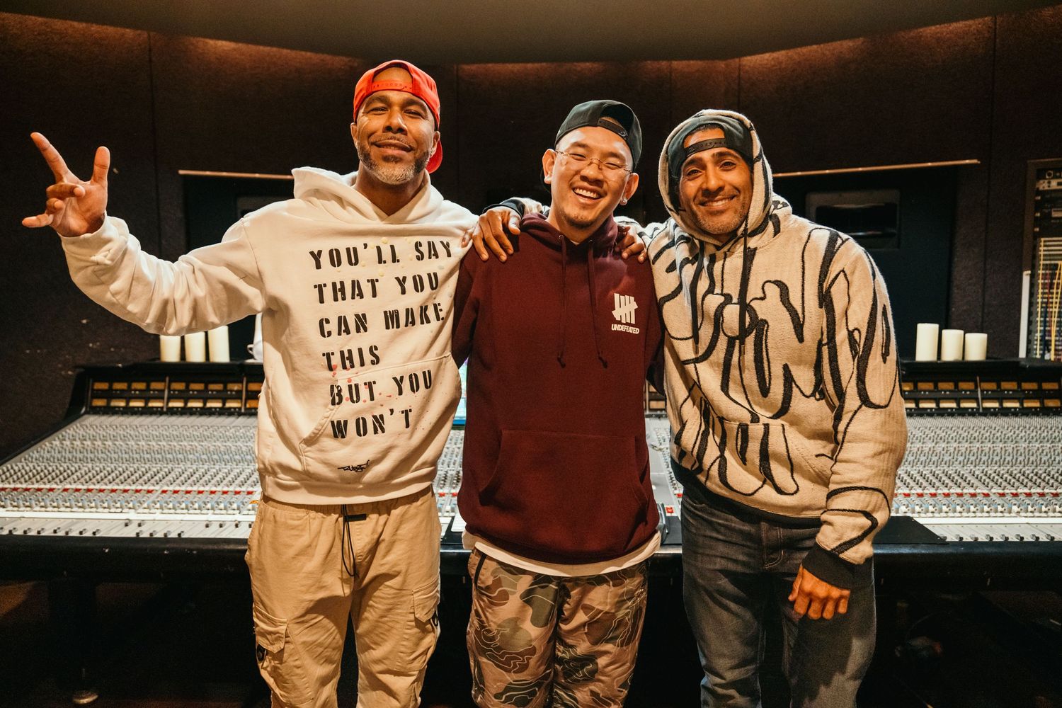 Three men smiling together in a recording studio with a large mixing console in the background.
