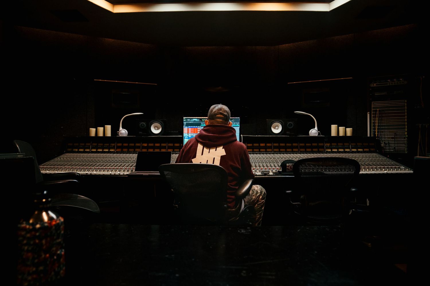 Music producer sitting alone at a large mixing console in a dark professional recording studio.
