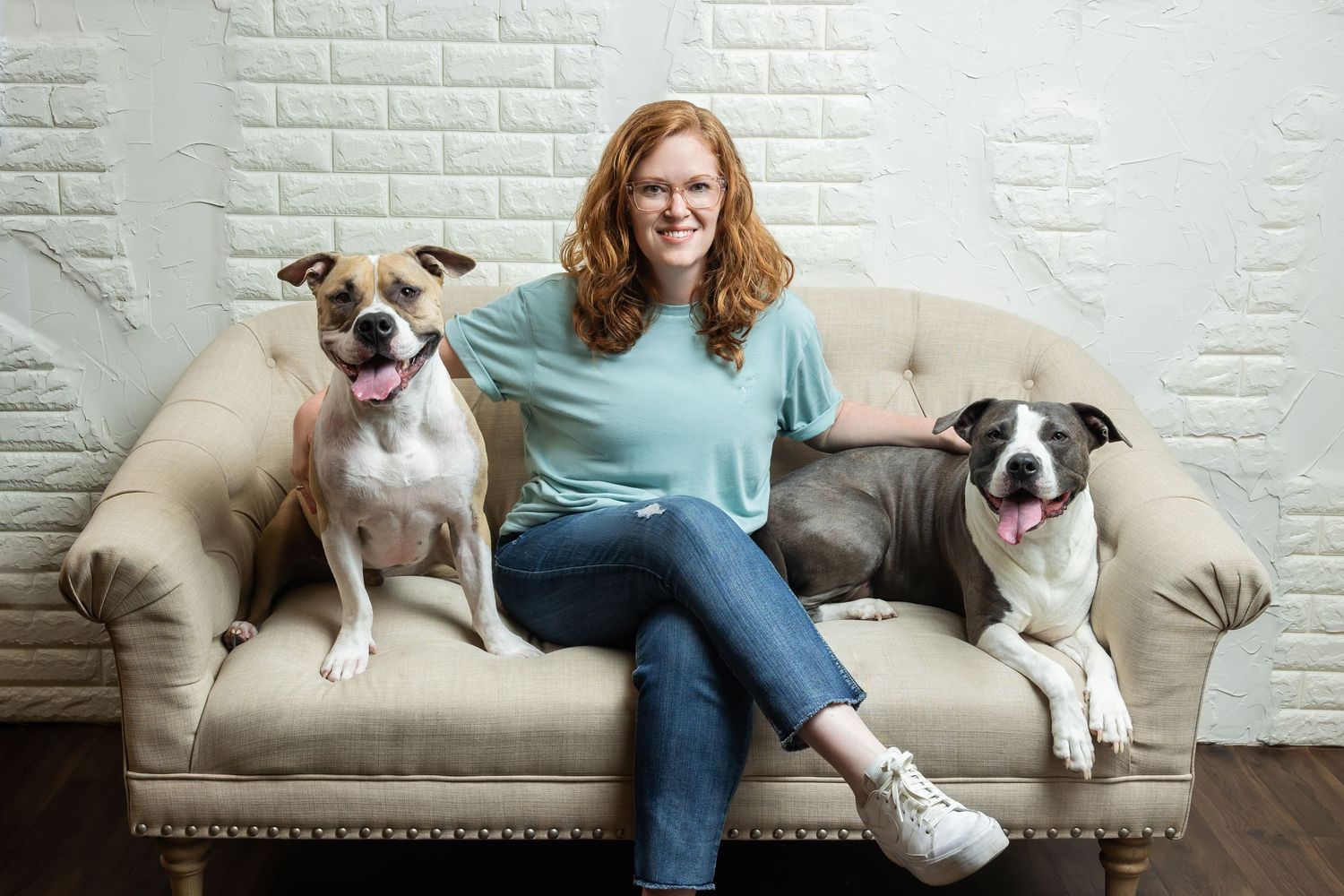 Woman with red hair sits on beige sofa between two happy pit bull dogs against white brick wall.