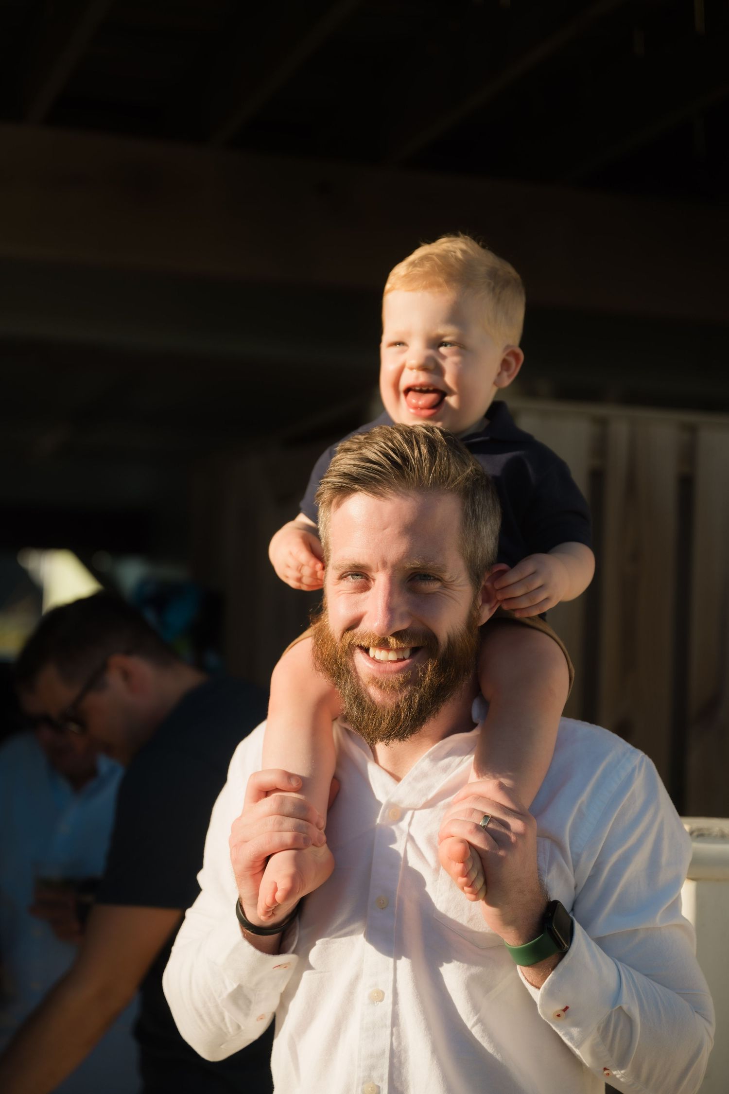 Smiling bearded man with toddler sitting on his shoulders, both laughing joyfully at an outdoor event.