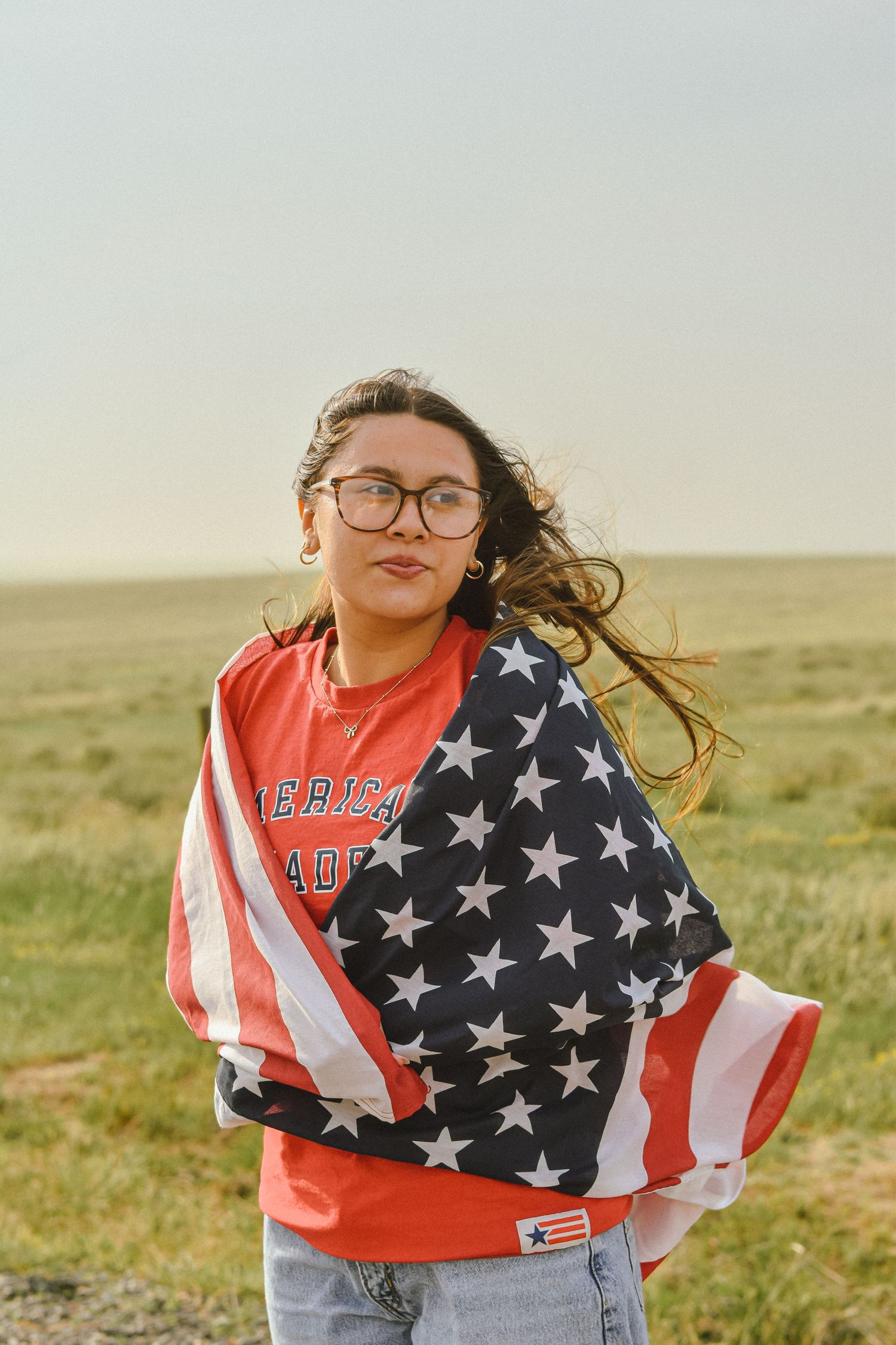 Young woman with glasses wrapped in an American flag, standing in a green field, wearing a red sweatshirt.